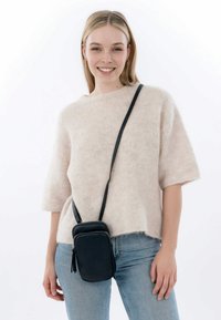 Black crossbody bag with a zippered closure and tassel detail, worn over a light pink, fuzzy knit sweater with short sleeves and denim jeans.