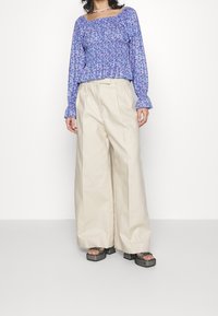 Bluse mit langen Ärmeln in blauem Blumenmuster und smocktem Taillenbund, kombiniert mit beigen weit geschnittenen Hosen und klobigen schwarzen Plateausandalen.
