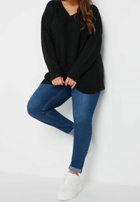 Kvinna i en svart oversize-tröja, blå uppvikta skinny jeans och vita sneakers, stående med ett ben böjt mot en vit vägg.