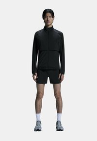 Sort letvægts atletisk jakke med lynlås foran, designet med en minimal æstetik. Matchet med sorte shorts og grå løbesko.