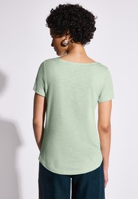 T-shirt s krátkým rukávem v mátově zelené barvě, s kulatým výstřihem a mírně zakřiveným lemováním. Vyrobený z měkkého, lehkého materiálu, s hladkým povrchem.