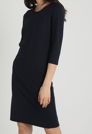 Femme portant une robe noire simple, longueur genou, manches trois-quarts sur un fond clair uni.