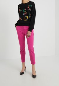 Woman wearing black sweatshirt with colorful floral letters, bright pink cropped pants, and black pointed-toe heels, standing against white background.
