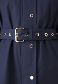 Impermeabil albastru marin cu talie accentuată prin curea, detalii din metal aurit, nasturi tip snapp și logo embosat pe țesătură. Textură netedă.