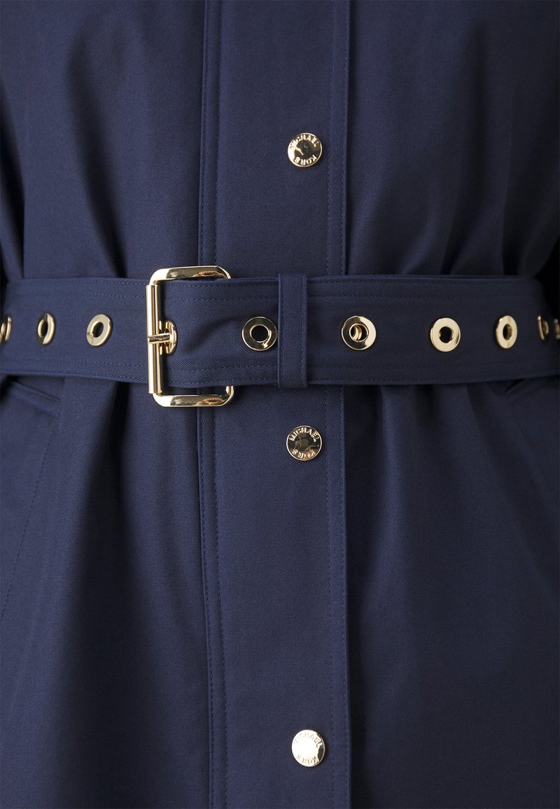 Chubasquero azul marino con cintura ceñida, herrajes dorados, botones a presión y detalle del logo en relieve en la tela. Textura suave.