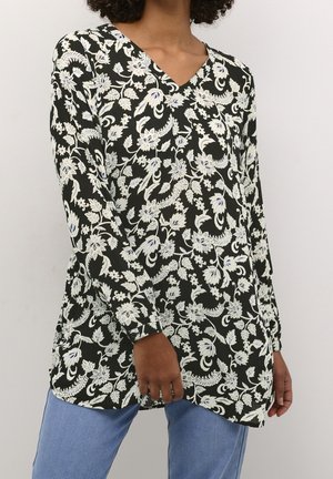 Femme portant une tunique noire à manches longues avec un col en V et un motif floral blanc, associée à un jean bleu clair, debout devant un fond uni.