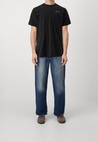 Camiseta negra de manga corta con escote redondo y un sutil detalle del logo en el pecho, combinada con unos vaqueros de denim ligeros y holgados de color azul. Zapatos marrones.
