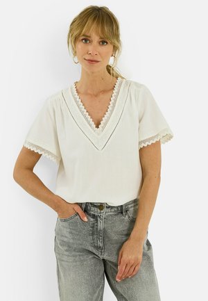 Femme aux cheveux blonds portant un chemisier blanc à col en V bordé de dentelle et un jean gris, debout devant un fond clair uni.