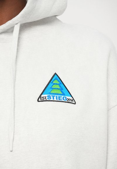 Svetlo siva kapuca s kengursko žepom. Ima trokutasti izvezeni pečat u plavoj i zelenoj boji, s tekstom "EST. STIEG 2014". Mekana tekstura.