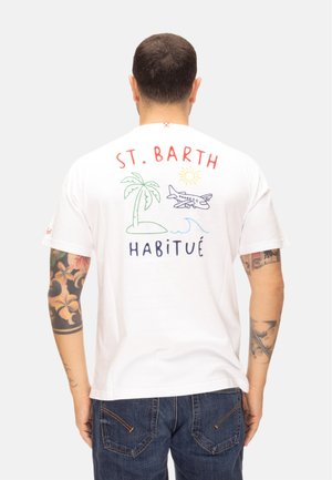 Uomo con tatuaggi indossa una maglietta bianca con un disegno colorato di St. Barth che mostra una palma, un aereo, il sole e onde oceaniche, abbinata a jeans blu.