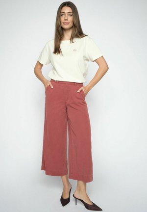 Frau steht vor neutralem Hintergrund, trägt ein cremefarbenes T-Shirt, weite, rostfarbene Hosen und dunkle spitz zulaufende Absätze, Hände in den Taschen.