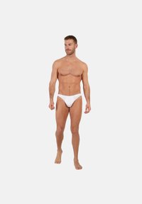 Uomo muscoloso che indossa mutande bianche da uomo, in piedi a piedi nudi contro uno sfondo semplice grigio chiaro, guardando leggermente verso destra.