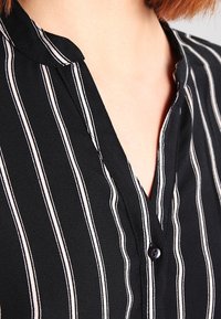 Chemise noire à col avec rayures blanches verticales ; dotée d'une patte de boutonnage et d'un tissu lisse et texturé.