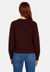 Kvinna med långt röd hår iklädd en mörk vinröd tröja och blå jeans, stående med ryggen mot en enkel ljus bakgrund.