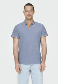 Camisa polo gris con cuello naranja y acentos de mangas a rayas, hecha de algodón, con un pequeño logo en el pecho. Llevada con jeans de color azul claro.