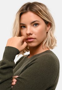 Olivgrön stickad tröja med rund halsringning och ribbade ärmslut, med mjuk textur och avspänd passform, bärs av en modell med ljust hår.