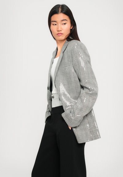Donna in giacca a scacchi grigia con sottili paillettes, top bianco e pantaloni neri a gamba larga, in piedi con le mani nelle tasche, che guarda di lato.