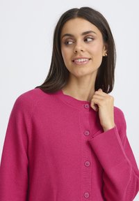 Cardigan tricotat roz, cu guler rotund, închidere centrală cu nasturi și manșete cu nervuri. Textură netedă, cu un model de împletire subtil.