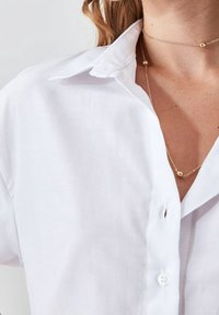 Camisa de algodón blanca con un collar clásico y cierre de botones. Una delicada cadena de oro con un acento de cuentas está superpuesta por debajo.