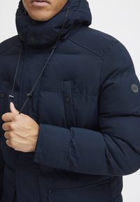 Navy puffer jacket with a high collar, drawstring hood, and two front pockets. Features snap buttons and a smooth, textured fabric.
