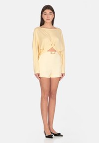 Top giallo a maniche lunghe con dettaglio annodato in vita, abbinato a pantaloni shorts a vita alta con un sottile accento in metallo dorato. Scarpe nere con punta.