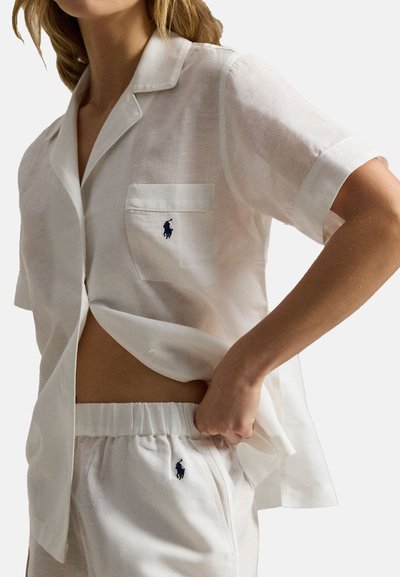 Vrouw draagt een wit kortärmelig overhemdd met knopen en bijpassende shorts met elastische tailleband, beide met kleine marineblauwe geborduurde logo's.