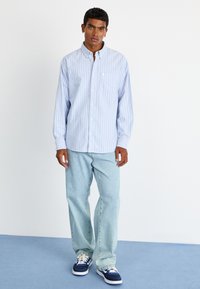 Light blue striped button-up shirt with a pocket, paired with light blue jeans and navy sneakers. Smooth fabric, relaxed fit.