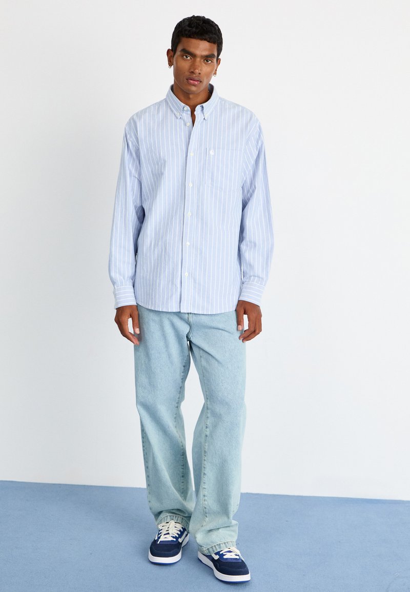 Chemise boutonnée rayée bleu clair avec une poche, assortie à un jean bleu clair et des baskets bleu marine. Tissu doux, coupe décontractée.