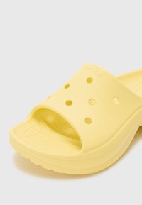 Dzeltena putu slaida ar atvērtu dizainu, kurā ir ventilācijas caurumi un strukturēts soļu laukums. Zīmola nosaukums "CROCS" ir iegravēts siksnā.