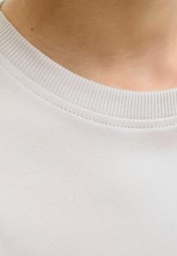 Lähedal vaade heledahalli tekstuuriga särkide kraele, näidates kudumisserva ja kaelapiirkonda, allpool on nähtav sile nahk.
