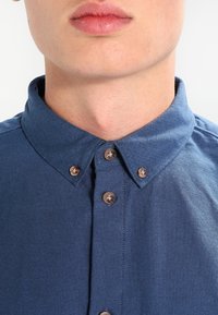 Blaues Hemd mit Button-Down-Kragen, braunen Knöpfen und strukturiertem Stoff. Fokus auf dem oberen Brustbereich ohne Hintergrundelemente.