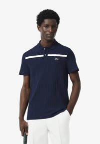 Polo de color azul marino hecho de tela acanalada, con una franja horizontal blanca y un logo verde de Lacoste en el pecho. Manga corta.