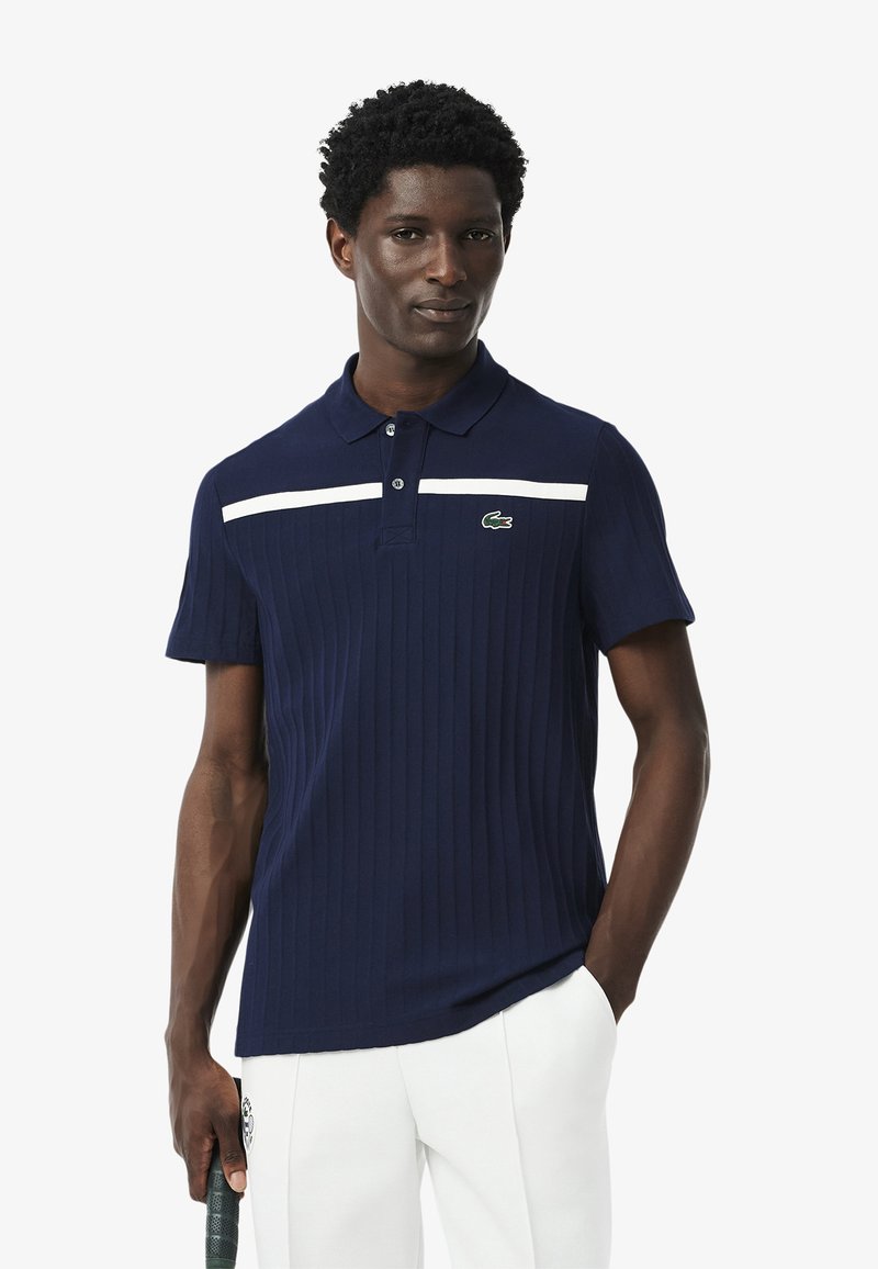Polo de color azul marino hecho de tela acanalada, con una franja horizontal blanca y un logo verde de Lacoste en el pecho. Manga corta.