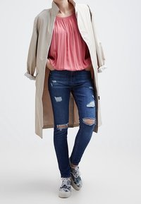 Beżowy płaszcz przeciwdeszczowy noszony na różowej bluzce w plisy, zestawiony z przetartymi niebieskimi dżinsami. Szare i białe, wzorzyste sneakersy dopełniają stylizację.