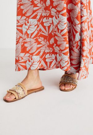 Pieds nus portant des sandales slip-on ornées, debout sur un sol blanc, avec une longue jupe orange présentant de grands motifs de feuilles blanches.