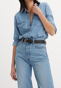 Femme portant une chemise en denim bleu clair à manches retroussées et un jean bleu clair taille haute maintenu par une ceinture en cuir noire avec une boucle argentée.
