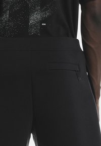 Pantaloni negri cu un material elegant, neted și cu un luciu subtil. Include un mic buzunar la spate cu închidere prin fermoar. Design minimalist.