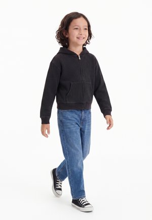 Lille barn går, iført en sort hættetrøje med lynlås, blå jeans og sorte sneakers med hvide snørebånd, smilende og kigger fremad.
