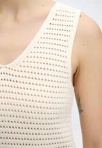 Κοντινό πλάνο ενός ατόμου που φοράει ένα κρεμ ρουχαλάκι πλεκτό, δείχνοντας ένα μικρό λουλουδένιο τατουάζ στον πάνω βραχίονα.