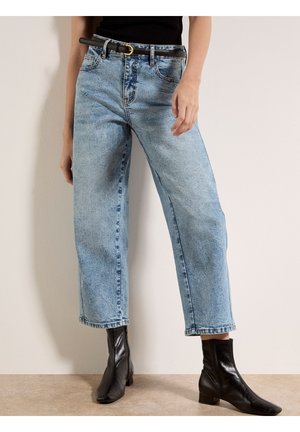 Donna che indossa jeans larghi cropped azzurri, cintura nera e stivaletti neri in piedi contro un muro beige.