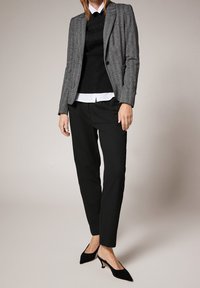 Veste en tweed gris par-dessus un pull noir et une chemise blanche, associée à un pantalon noir fuselé et des chaussures à petit talon noir.