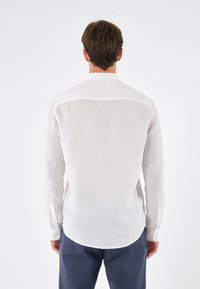 Uomo con capelli corti castani, vestito con una camicia bianca a maniche lunghe e pantaloni scuri, in piedi di spalle contro uno sfondo bianco uniforme.