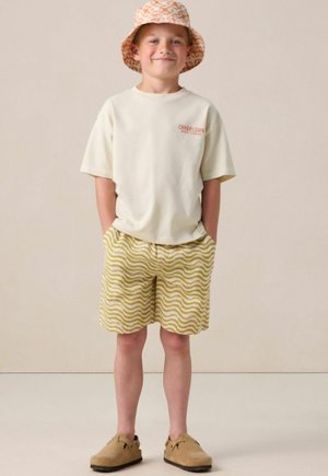 Smiling boy wearing a patterned bucket hat, white short-sleeve shirt, yellow wavy striped shorts, and tan slip-on shoes, standing with hands in pockets.