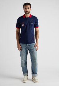 Granatowa koszulka polo z czerwonym kołnierzykiem i rękawami, z zamkiem błyskawicznym, naszywkami haftowanymi oraz jasnoniebieskimi dżinsami w połączeniu z beżowymi sneakersami.