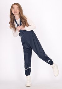 Šypsena mergaitė su ilgais plaukais, apsivilkusi jūros mėlynos spalvos vandeniui nepralaidžius kombinezonus ir baltus sportinius batelius, pakėlusi vieną koją priešais vientisą baltą foną.