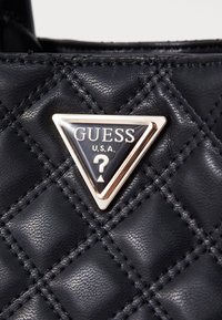 Sort quiltet syntetisk læder med et trekantet guld-accenteret mærke, der viser "GUESS U.S.A." og et dekorativt spørgsmålstegn emblem.