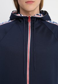 Mörkblå zip hoodie med fodrad huva, röd dragkedja, vit accent och blå- och vitrandiga axeldetaljer. Slätt, stretchigt tyg.