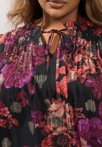 Blusa nera con vivaci motivi floreali in rosso, viola e oro. Presenta un colletto arricciato e una parte centrale con un nastro da legare davanti.