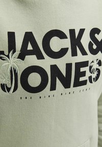 Texte noir "JACK & JONES" sur tissu clair avec des palmiers brodés à l'intérieur des lettres J et S, et petit texte "ONE MINE NINE ZERO" en dessous.