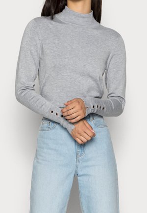 Woman wearing light grey long-sleeve turtleneck sweater with button details on cuffs and light blue high-waisted jeans.